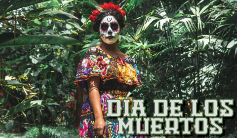 A woman in traditional Dia de los muertos make up and outfit surrounded by tropical leaves